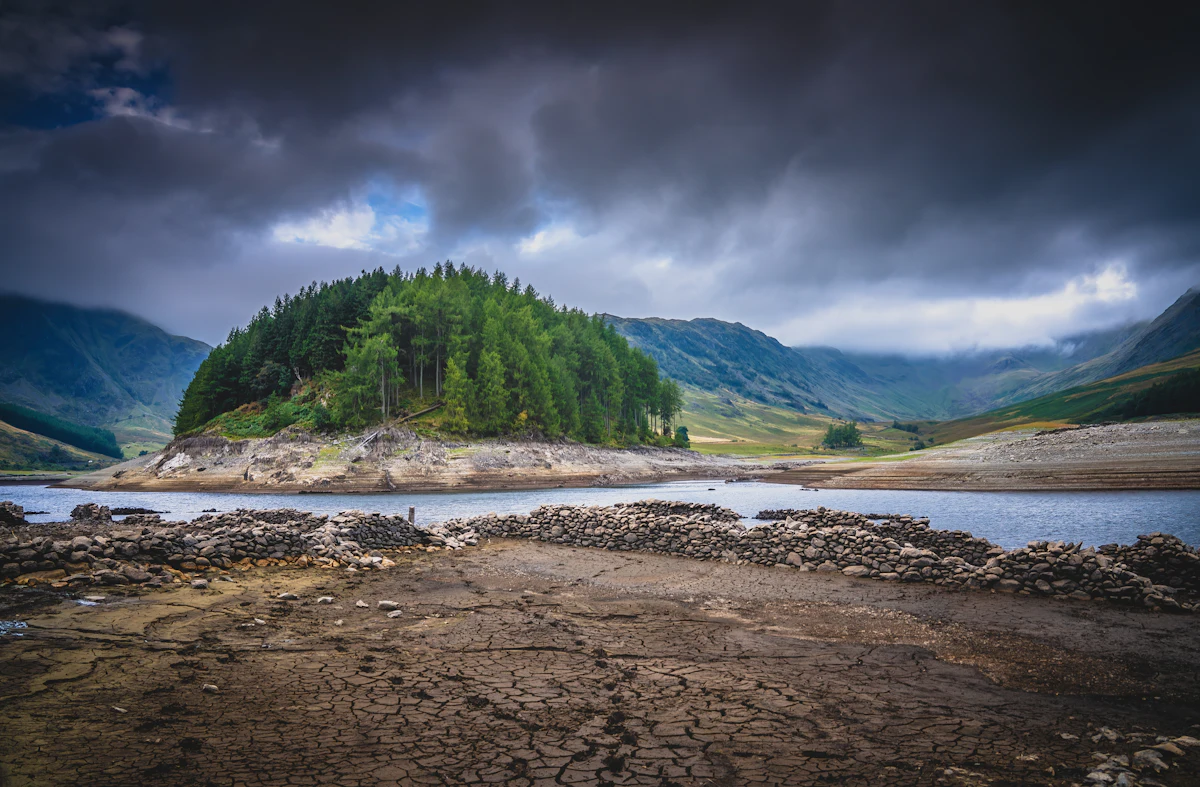 A wooded island and a dry, cracked shoreline at Haweswater Reservoir under dark cloud