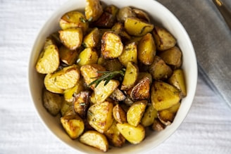 A bowl filled with golden, roasted potato pieces garnished with rosemary. The potatoes have a crispy appearance with varying shades of brown, indicating they are well-cooked. The background is a textured, light-colored fabric.