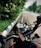 A motorcycle's dashboard is prominently displayed with a keychain attached. The background reveals a wet, deserted road lined with green trees and a blue-and-white-striped curb.