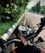 A motorcycle's dashboard is prominently displayed with a keychain attached. The background reveals a wet, deserted road lined with green trees and a blue-and-white-striped curb.