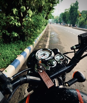 A motorcycle's dashboard is prominently displayed with a keychain attached. The background reveals a wet, deserted road lined with green trees and a blue-and-white-striped curb.
