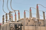 An array of electrical insulators and conductors on top of a large power transformer. The insulators have a cylindrical, ribbed design to prevent electrical leaks, and there are several wires and cables connected to them. Cooling fans are visible on the side of the transformer.