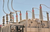 An array of electrical insulators and conductors on top of a large power transformer. The insulators have a cylindrical, ribbed design to prevent electrical leaks, and there are several wires and cables connected to them. Cooling fans are visible on the side of the transformer.