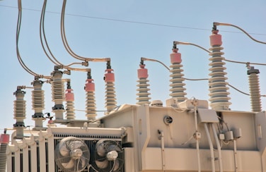 An array of electrical insulators and conductors on top of a large power transformer. The insulators have a cylindrical, ribbed design to prevent electrical leaks, and there are several wires and cables connected to them. Cooling fans are visible on the side of the transformer.