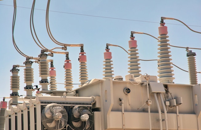 An array of electrical insulators and conductors on top of a large power transformer. The insulators have a cylindrical, ribbed design to prevent electrical leaks, and there are several wires and cables connected to them. Cooling fans are visible on the side of the transformer.
