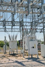 An industrial power substation with a complex network of transformers and electrical components interconnected by various wires and metal structures. The scene is composed of metal towers, insulators, and other electrical equipment situated on a gravel-covered ground, with some greenery visible in the background.