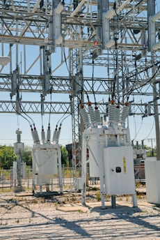 An industrial power substation with a complex network of transformers and electrical components interconnected by various wires and metal structures. The scene is composed of metal towers, insulators, and other electrical equipment situated on a gravel-covered ground, with some greenery visible in the background.