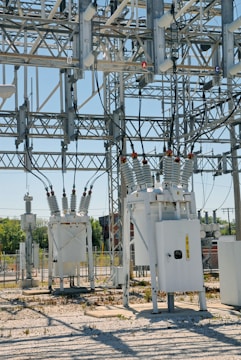 An industrial power substation with a complex network of transformers and electrical components interconnected by various wires and metal structures. The scene is composed of metal towers, insulators, and other electrical equipment situated on a gravel-covered ground, with some greenery visible in the background.