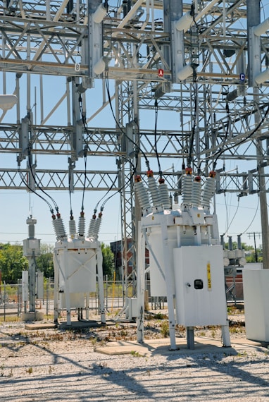 An industrial power substation with a complex network of transformers and electrical components interconnected by various wires and metal structures. The scene is composed of metal towers, insulators, and other electrical equipment situated on a gravel-covered ground, with some greenery visible in the background.