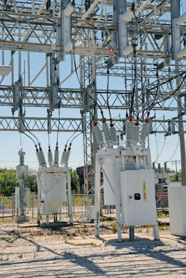 An industrial power substation with a complex network of transformers and electrical components interconnected by various wires and metal structures. The scene is composed of metal towers, insulators, and other electrical equipment situated on a gravel-covered ground, with some greenery visible in the background.