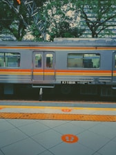 A train with a silver and orange exterior is parked at a station platform. The train has several windows, and there's greenery, including trees, in the background. The platform has tiled flooring marked with bright orange symbols and tactile paving, indicating waiting areas.