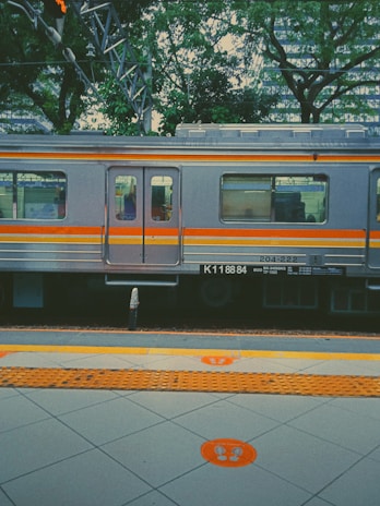 A train with a silver and orange exterior is parked at a station platform. The train has several windows, and there's greenery, including trees, in the background. The platform has tiled flooring marked with bright orange symbols and tactile paving, indicating waiting areas.