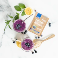 two glasses of blueberry smoothie next to a bag of protein