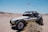 A dune buggy is parked on a sandy desert landscape. The vehicle has a frame with visible seats, large off-road tires, and a red flag attached to a tall antenna. In the background, there are arid hills and a few scattered bushes.