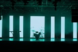 A dimly lit stage features a set of musical instruments including a drum kit and a keyboard. Behind the instruments, large vertical LED panels emit a turquoise glow, creating a modern and atmospheric backdrop. The ceiling is equipped with various stage lights and rigging.