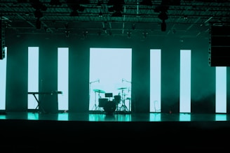 A dimly lit stage features a set of musical instruments including a drum kit and a keyboard. Behind the instruments, large vertical LED panels emit a turquoise glow, creating a modern and atmospheric backdrop. The ceiling is equipped with various stage lights and rigging.