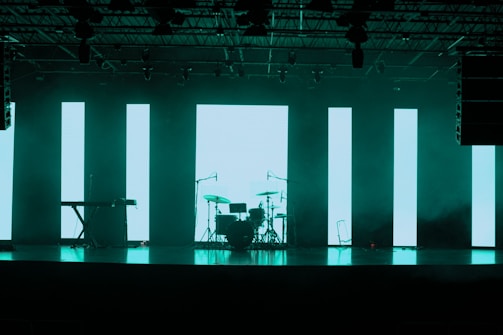 A dimly lit stage features a set of musical instruments including a drum kit and a keyboard. Behind the instruments, large vertical LED panels emit a turquoise glow, creating a modern and atmospheric backdrop. The ceiling is equipped with various stage lights and rigging.