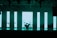 A dimly lit stage features a set of musical instruments including a drum kit and a keyboard. Behind the instruments, large vertical LED panels emit a turquoise glow, creating a modern and atmospheric backdrop. The ceiling is equipped with various stage lights and rigging.