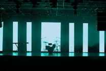A dimly lit stage features a set of musical instruments including a drum kit and a keyboard. Behind the instruments, large vertical LED panels emit a turquoise glow, creating a modern and atmospheric backdrop. The ceiling is equipped with various stage lights and rigging.