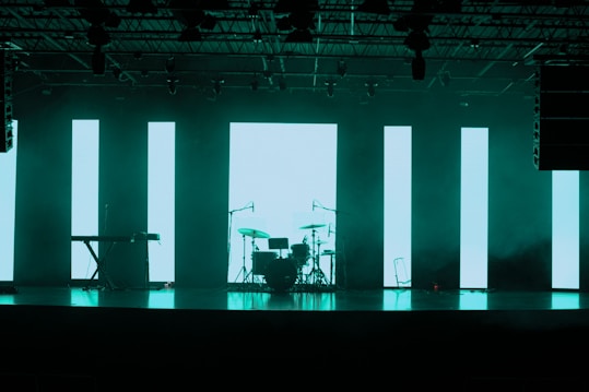 A dimly lit stage features a set of musical instruments including a drum kit and a keyboard. Behind the instruments, large vertical LED panels emit a turquoise glow, creating a modern and atmospheric backdrop. The ceiling is equipped with various stage lights and rigging.