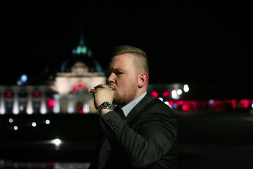A man with short hair and wearing a suit stands outdoors at night, holding a cigar near his mouth. The background features a large, illuminated building with decorative lights, creating a luxurious and sophisticated atmosphere.