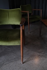 Two vintage green upholstered chairs with wooden arms and legs are positioned on a concrete floor. The scene is dimly lit, creating a cozy and classic ambiance.