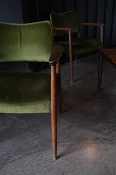 Two vintage green upholstered chairs with wooden arms and legs are positioned on a concrete floor. The scene is dimly lit, creating a cozy and classic ambiance.