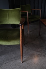 Two vintage green upholstered chairs with wooden arms and legs are positioned on a concrete floor. The scene is dimly lit, creating a cozy and classic ambiance.