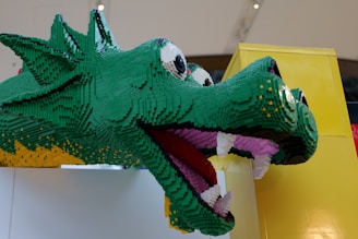 A large dragon head constructed from green, yellow, black, and purple LEGO bricks. The dragon has big eyes with a playful expression, exhibiting white teeth and a pink tongue. It is mounted against a yellow structure, creating a vibrant and whimsical appearance.