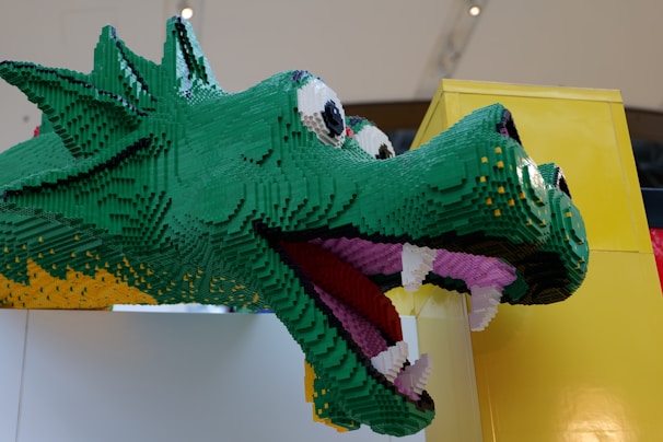 A large dragon head constructed from green, yellow, black, and purple LEGO bricks. The dragon has big eyes with a playful expression, exhibiting white teeth and a pink tongue. It is mounted against a yellow structure, creating a vibrant and whimsical appearance.