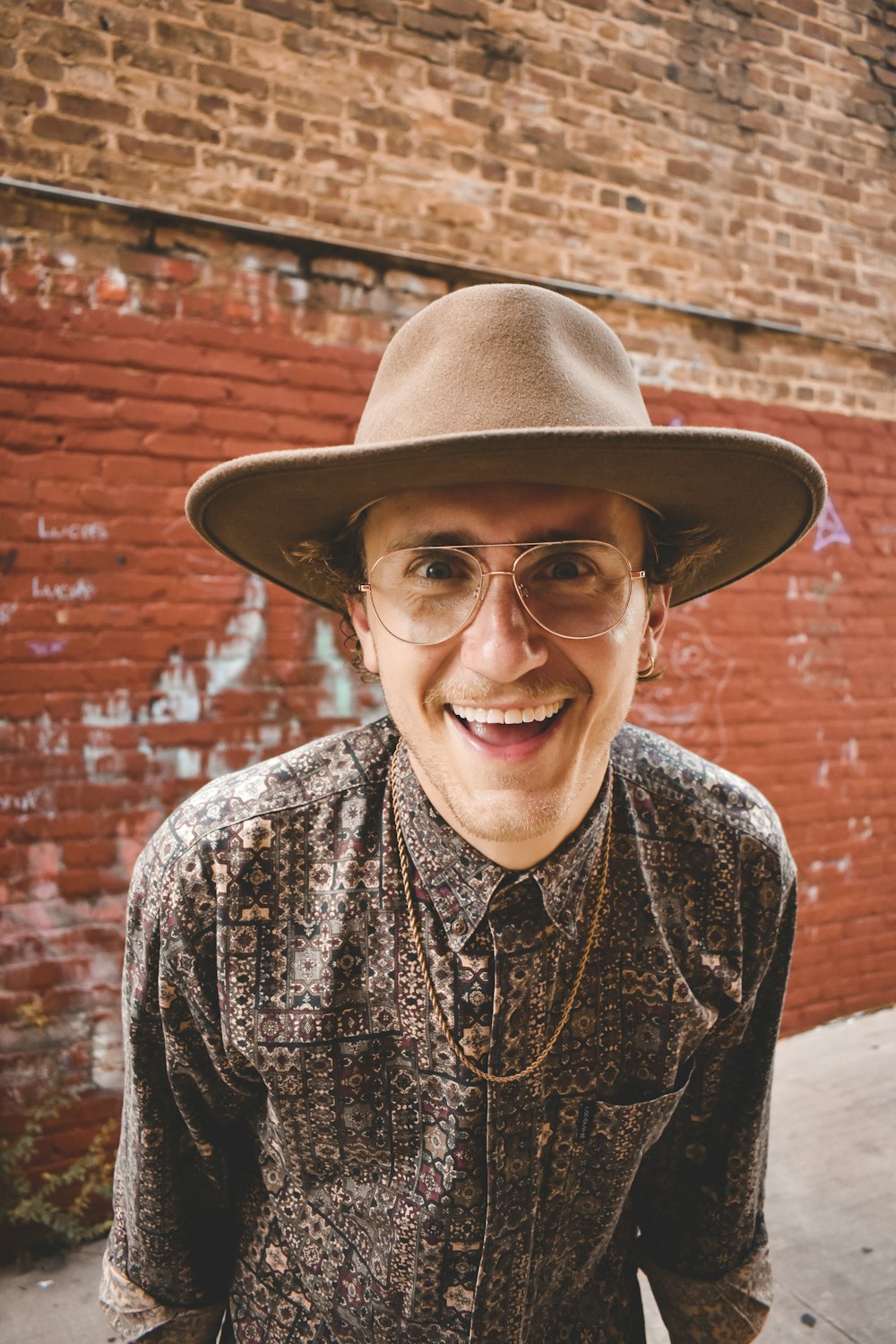 a man wearing a hat and glasses standing in front of a brick wall