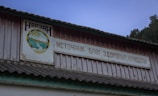 A weathered building facade features a wooden sign written in Russian with an emblem depicting mountains and a flowing river. The structure has a corrugated roof, and a backdrop of trees and a dusk sky.