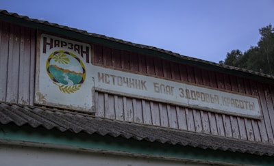 A weathered building facade features a wooden sign written in Russian with an emblem depicting mountains and a flowing river. The structure has a corrugated roof, and a backdrop of trees and a dusk sky.