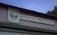 A weathered building facade features a wooden sign written in Russian with an emblem depicting mountains and a flowing river. The structure has a corrugated roof, and a backdrop of trees and a dusk sky.
