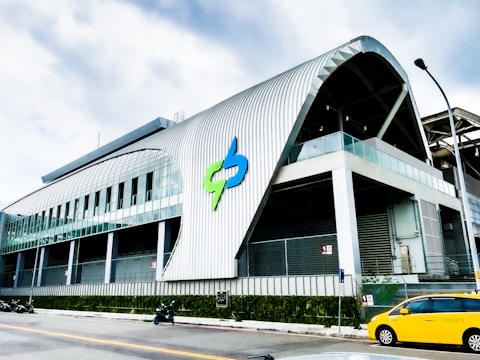 A modern, industrial building with a large metallic exterior featuring a curved design. The structure has multiple windows and a prominent logo on the facade. There are a few parked motorcycles and a yellow taxi visible on the street in front.