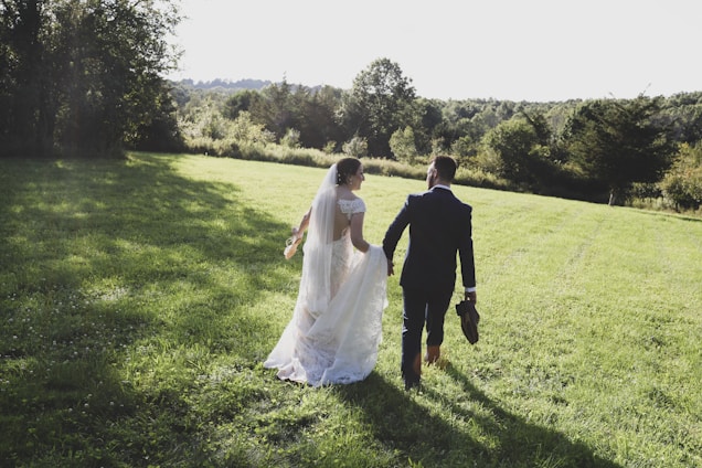 A bride and groom walk hand in hand across a lush, sunlit field. The bride wears a flowing white gown with lace detail and a veil, while the groom is dressed in a dark suit and carries his shoes. The scene is set in a serene natural landscape with trees and greenery in the background under a clear sky.