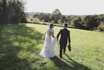 A bride and groom walk hand in hand across a lush, sunlit field. The bride wears a flowing white gown with lace detail and a veil, while the groom is dressed in a dark suit and carries his shoes. The scene is set in a serene natural landscape with trees and greenery in the background under a clear sky.