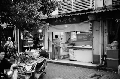 A bustling street market scene in black and white featuring a small shop with signs in Chinese. A person stands outside the shop, looking at a phone, while another person works inside attending to produce. Several bicycles and a motorbike are parked in the vicinity, and other individuals are moving through the scene under the shade of trees.