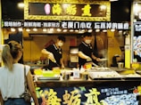 A street food vendor is preparing and grilling seafood at a food stall in an indoor market. The vendor is wearing a mask and uniform, focusing on cooking multiple skewers over an open grill. The stall is decorated with Chinese characters and lit by multiple overhead lights. A customer stands in front, observing the cooking process.
