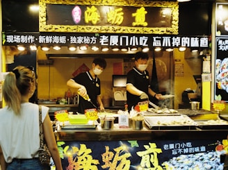 A street food vendor is preparing and grilling seafood at a food stall in an indoor market. The vendor is wearing a mask and uniform, focusing on cooking multiple skewers over an open grill. The stall is decorated with Chinese characters and lit by multiple overhead lights. A customer stands in front, observing the cooking process.