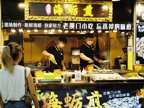 A street food vendor is preparing and grilling seafood at a food stall in an indoor market. The vendor is wearing a mask and uniform, focusing on cooking multiple skewers over an open grill. The stall is decorated with Chinese characters and lit by multiple overhead lights. A customer stands in front, observing the cooking process.