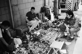 A street market scene featuring three individuals interacting with a variety of objects displayed on the ground. The items include trinkets, small sculptures, and various artifacts, organized in rows. The setting appears to be urban with a brick wall and a narrow alleyway.