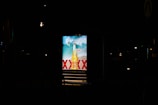 A brightly lit advertisement display at night featuring a beer bottle with waves in the background and the word 'NEW' prominently displayed. The letters 'XXXX' are visible, indicating a brand name.