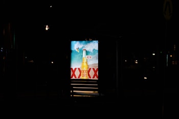 A brightly lit advertisement display at night featuring a beer bottle with waves in the background and the word 'NEW' prominently displayed. The letters 'XXXX' are visible, indicating a brand name.
