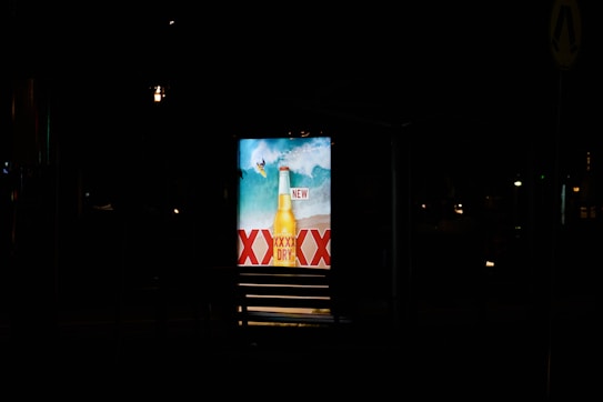 A brightly lit advertisement display at night featuring a beer bottle with waves in the background and the word 'NEW' prominently displayed. The letters 'XXXX' are visible, indicating a brand name.