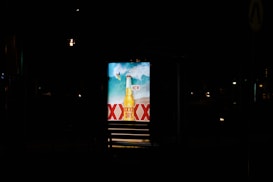 A brightly lit advertisement display at night featuring a beer bottle with waves in the background and the word 'NEW' prominently displayed. The letters 'XXXX' are visible, indicating a brand name.