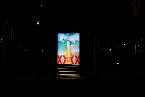 A brightly lit advertisement display at night featuring a beer bottle with waves in the background and the word 'NEW' prominently displayed. The letters 'XXXX' are visible, indicating a brand name.