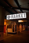 An illuminated 'MARKET' sign hangs above a sidewalk at night. The environment has dim lighting with various shop fronts visible, including one marked as a meat store. There are colorful handwritten signs on display, possibly detailing products or offers.