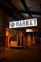 An illuminated 'MARKET' sign hangs above a sidewalk at night. The environment has dim lighting with various shop fronts visible, including one marked as a meat store. There are colorful handwritten signs on display, possibly detailing products or offers.