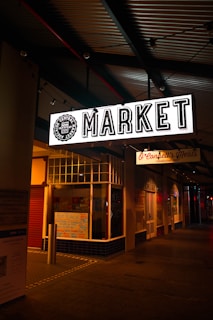 An illuminated 'MARKET' sign hangs above a sidewalk at night. The environment has dim lighting with various shop fronts visible, including one marked as a meat store. There are colorful handwritten signs on display, possibly detailing products or offers.